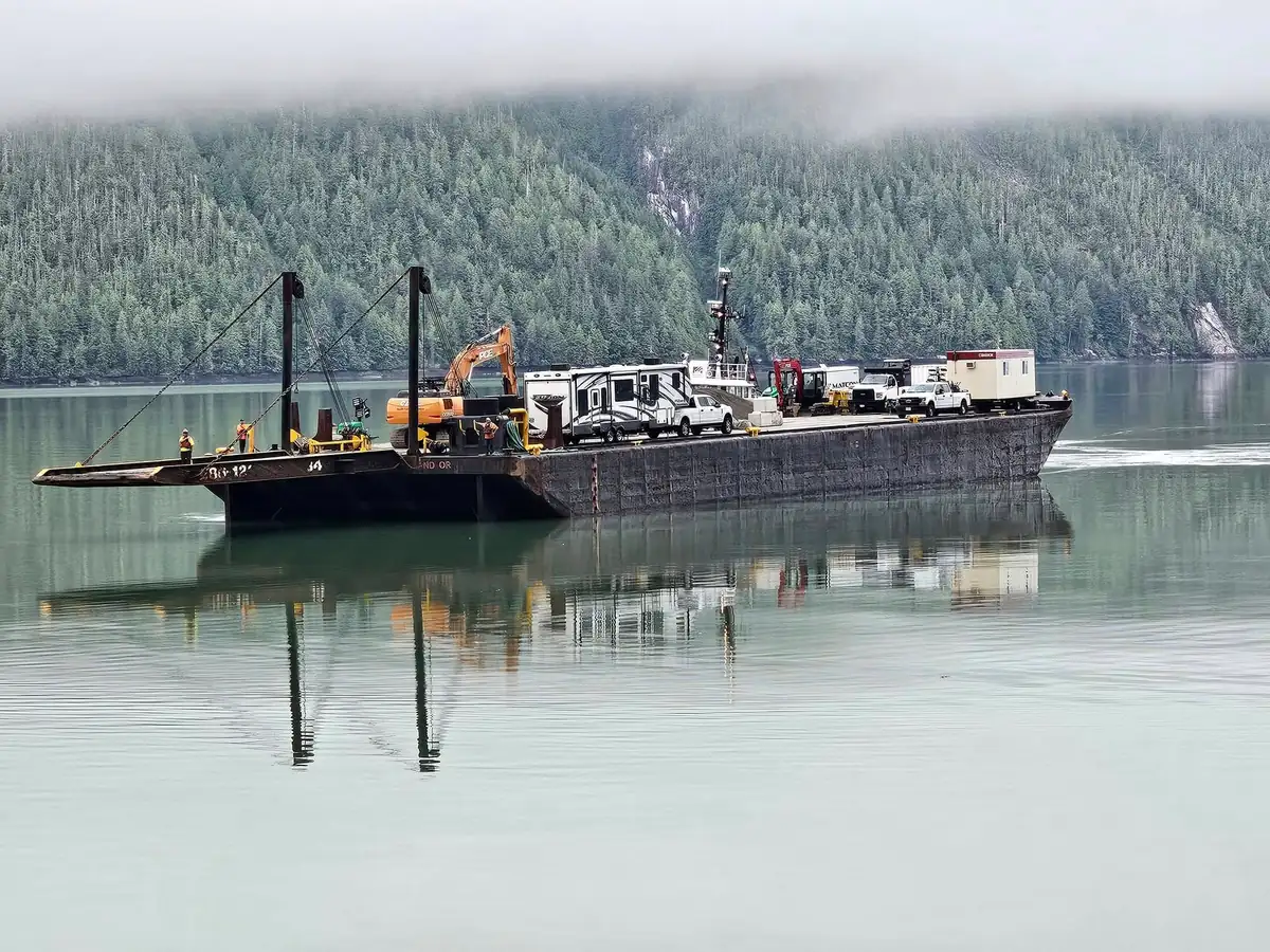Barge with PCE excavator, RV and pickup crossing the inlet under low cloud